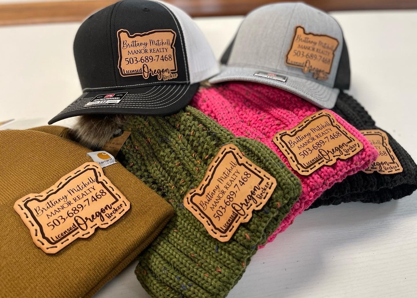 Collection of trucker hats and knit beanies with Oregon shaped leather patches for Manor Realty
